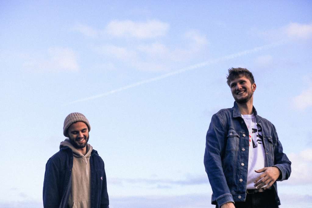 Tom and Greg in denim jackets candidly smiling with a blue sky behind them.